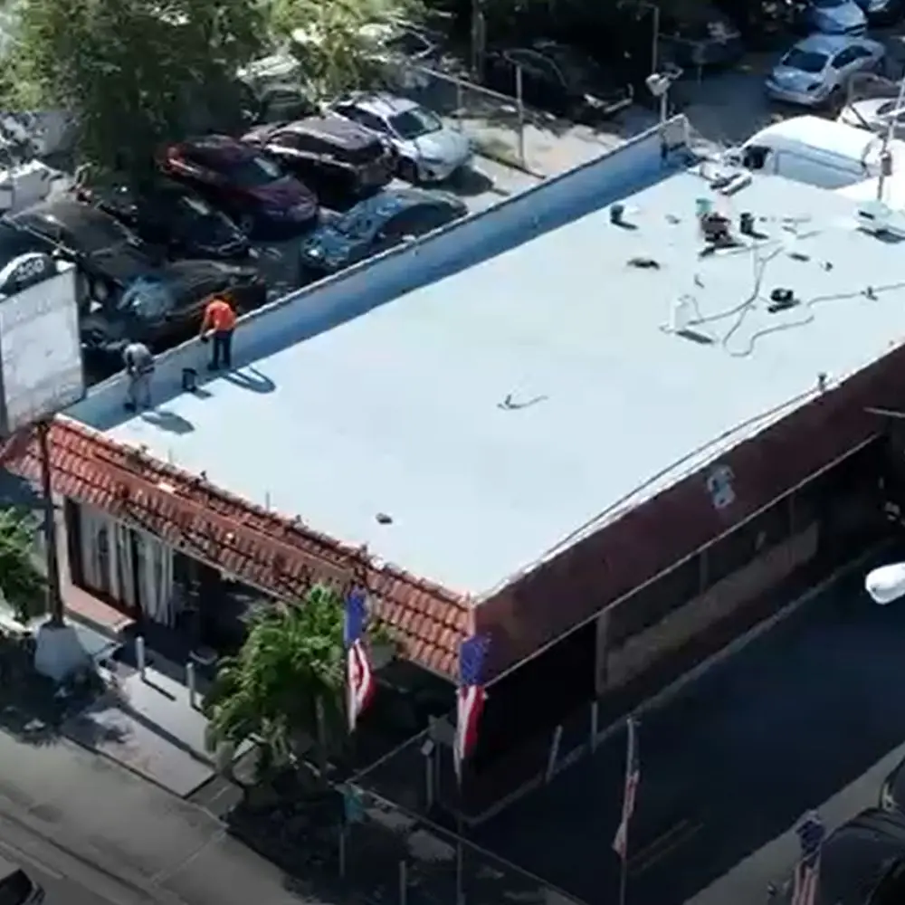 Aerial view of a commercial building undergoing TPO roof installation, with workers on the rooftop, surrounded by parked cars and a vibrant urban setting. Highlights the durability and energy efficiency of TPO roofing systems.