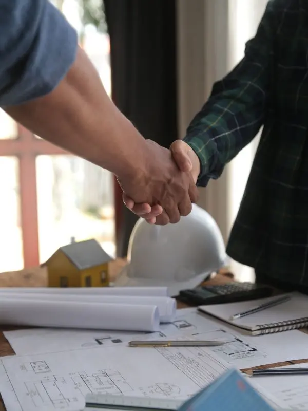 Two individuals shaking hands over a table with architectural plans, a hard hat, and construction tools, symbolizing a successful partnership. Represents professional services offered by Hernandez Morrice Services Inc.