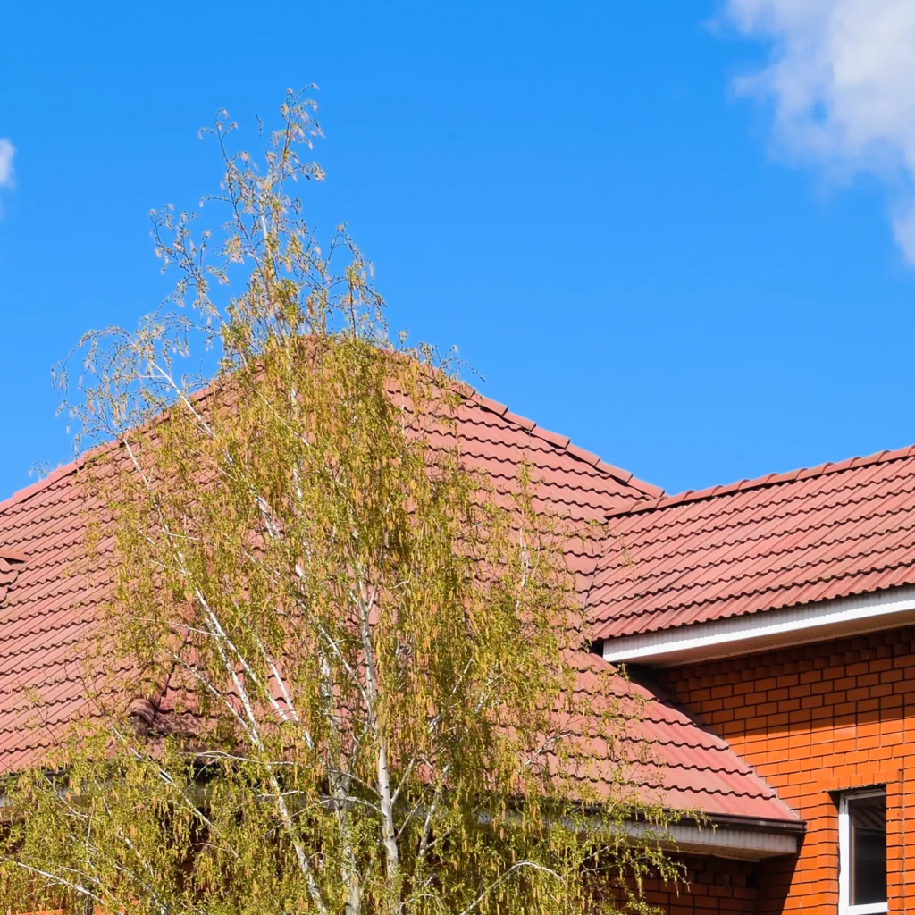 Bright red tile roof on a brick house with a tree in the foreground and a clear blue sky in the background. Quality roofing services by Hernandez Morrice Services Inc.