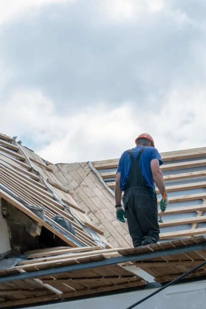 A construction worker in a blue shirt and protective gear standing on a roof under repair, showing wooden beams and an unfinished roof structure during a roof replacement project.